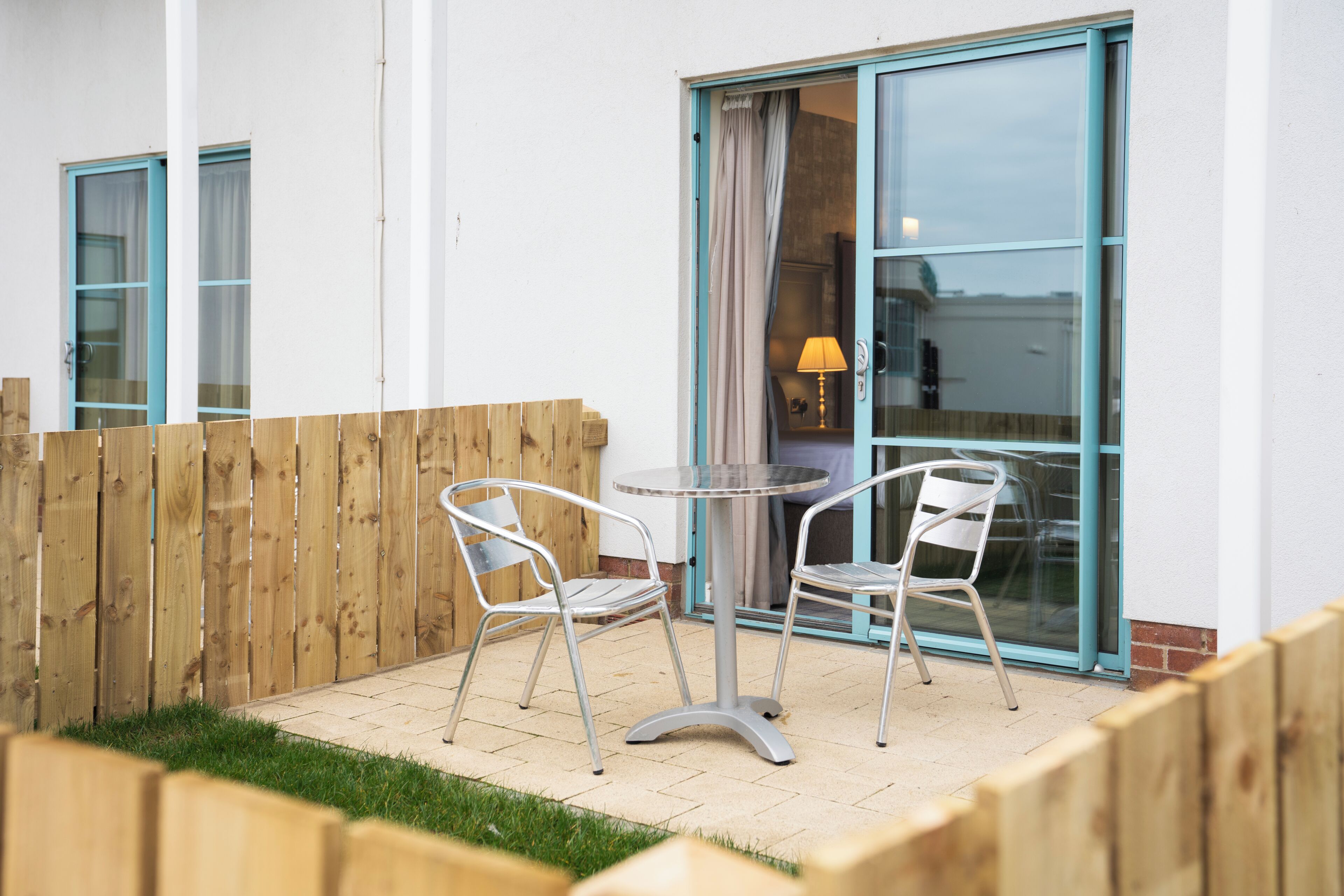 Une terrasse avec deux chaises en métal et une table, une clôture en bois et une porte-fenêtre coulissante avec un rideau.