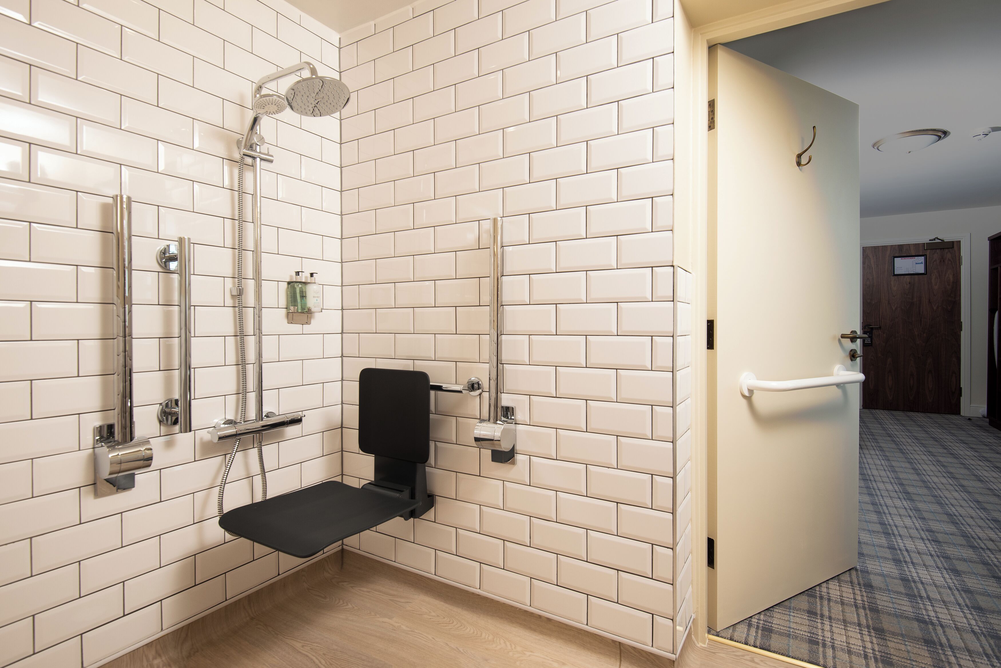 A bathroom with white tiled walls, a shower with a handheld showerhead, a toilet with a grab bar, and a door with a towel rack.