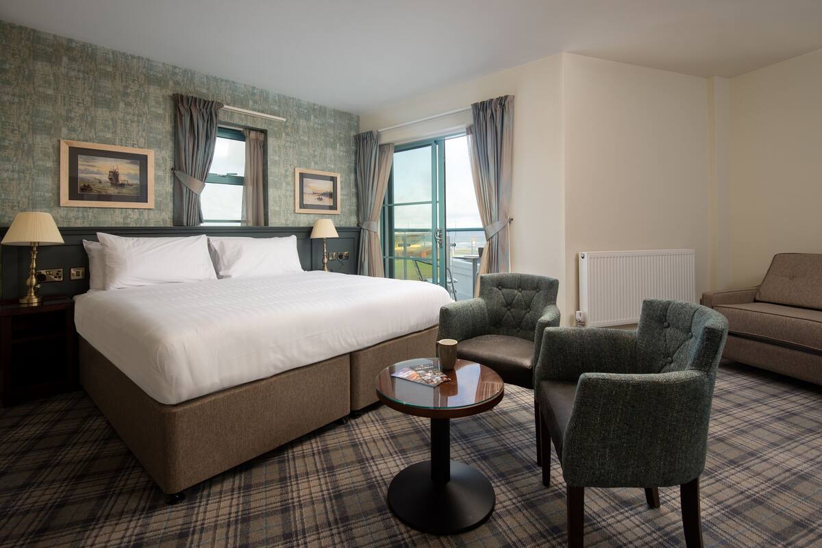 A hotel room with a large bed, two armchairs, a small round table, and a view of the outdoors through a window.