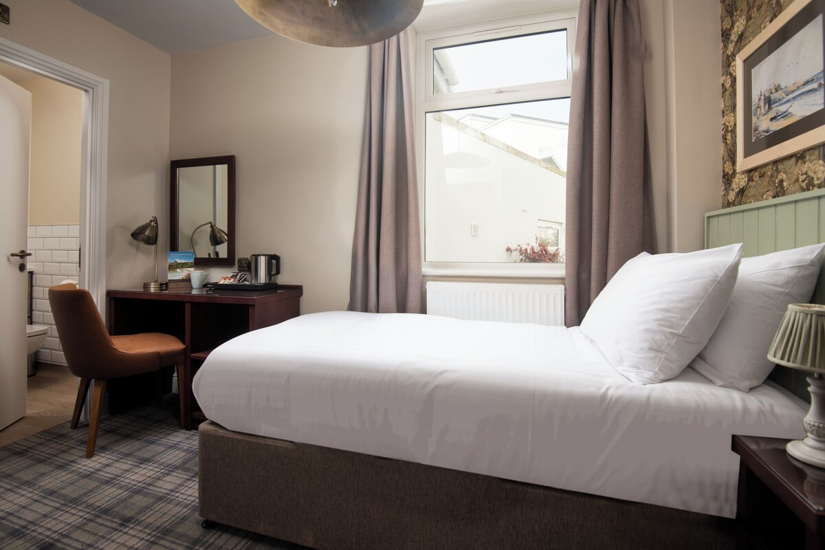 A hotel room with a bed, a desk, a chair, a window with curtains, and a painting on the wall.