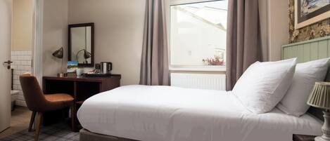 A hotel room with a bed, a desk, a chair, a window with curtains, and a painting on the wall.