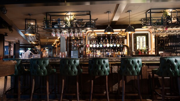 A bar area with wooden flooring, green tufted bar stools, and a variety of bottles and glasses on shelves. - The Tynemouth Castle Inn - The Inn Collection Group (North Shields)