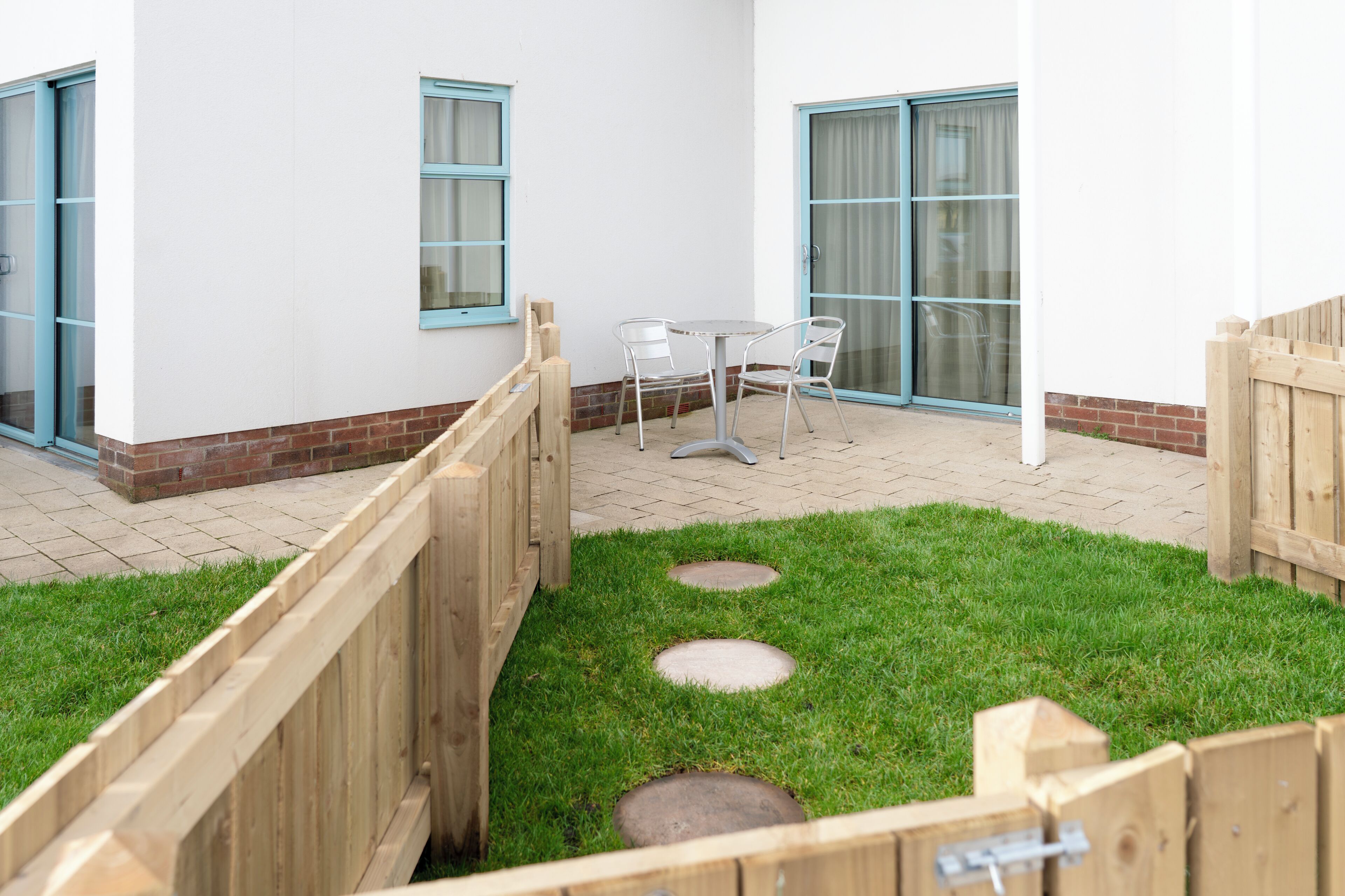 A patio area with a table and chairs, a wooden fence, and a grassy lawn with stepping stones.