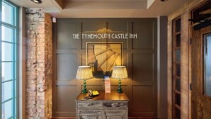 A reception area with a sign reading 'The Tynewyth Castle Inn,' a wooden desk with two lamps, and a brick wall. - The Tynemouth Castle Inn - The Inn Collection Group (North Shields)