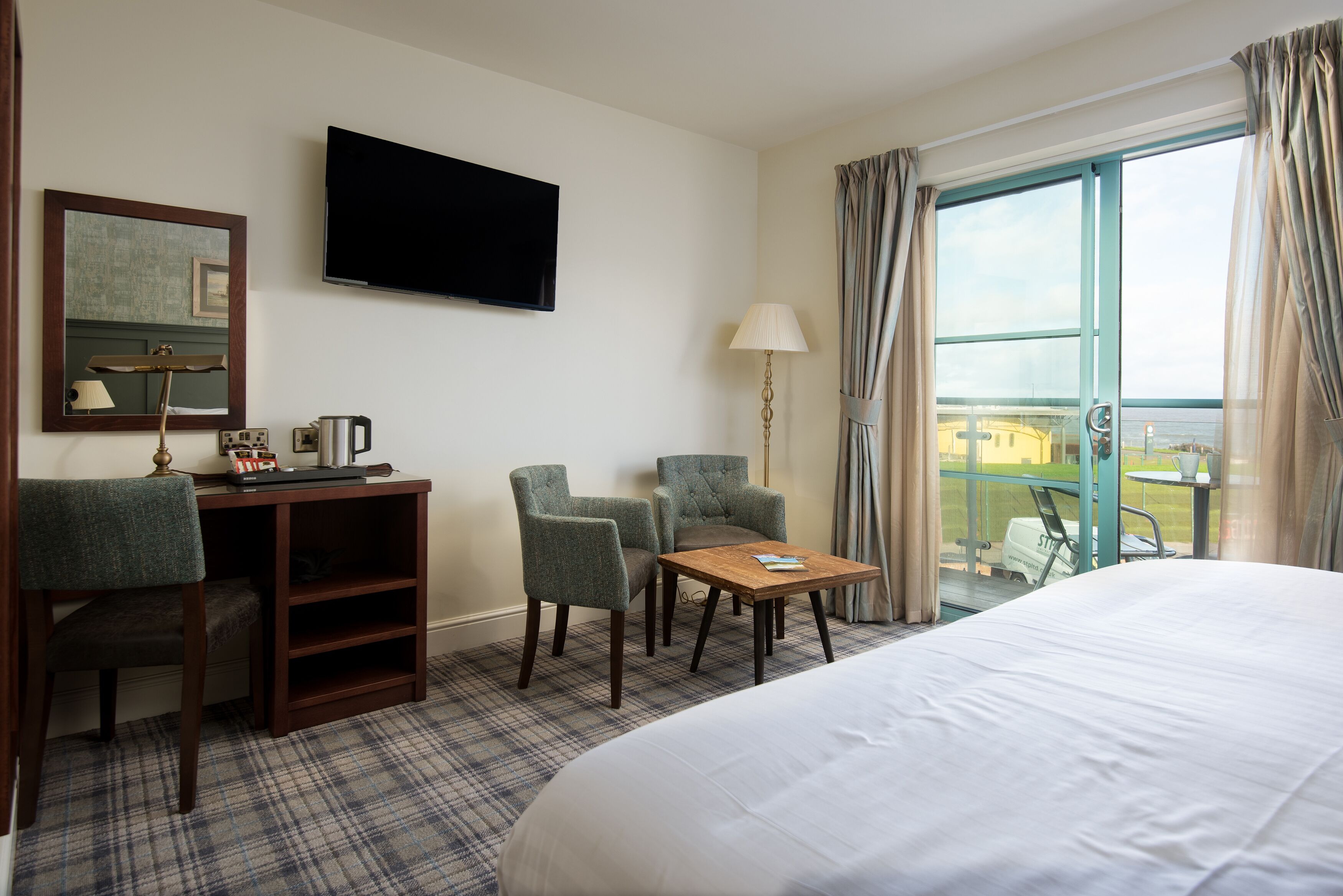 Habitación de hotel con cama, dos sillones, una mesita, televisor, escritorio con lámpara y ventana con cortinas.