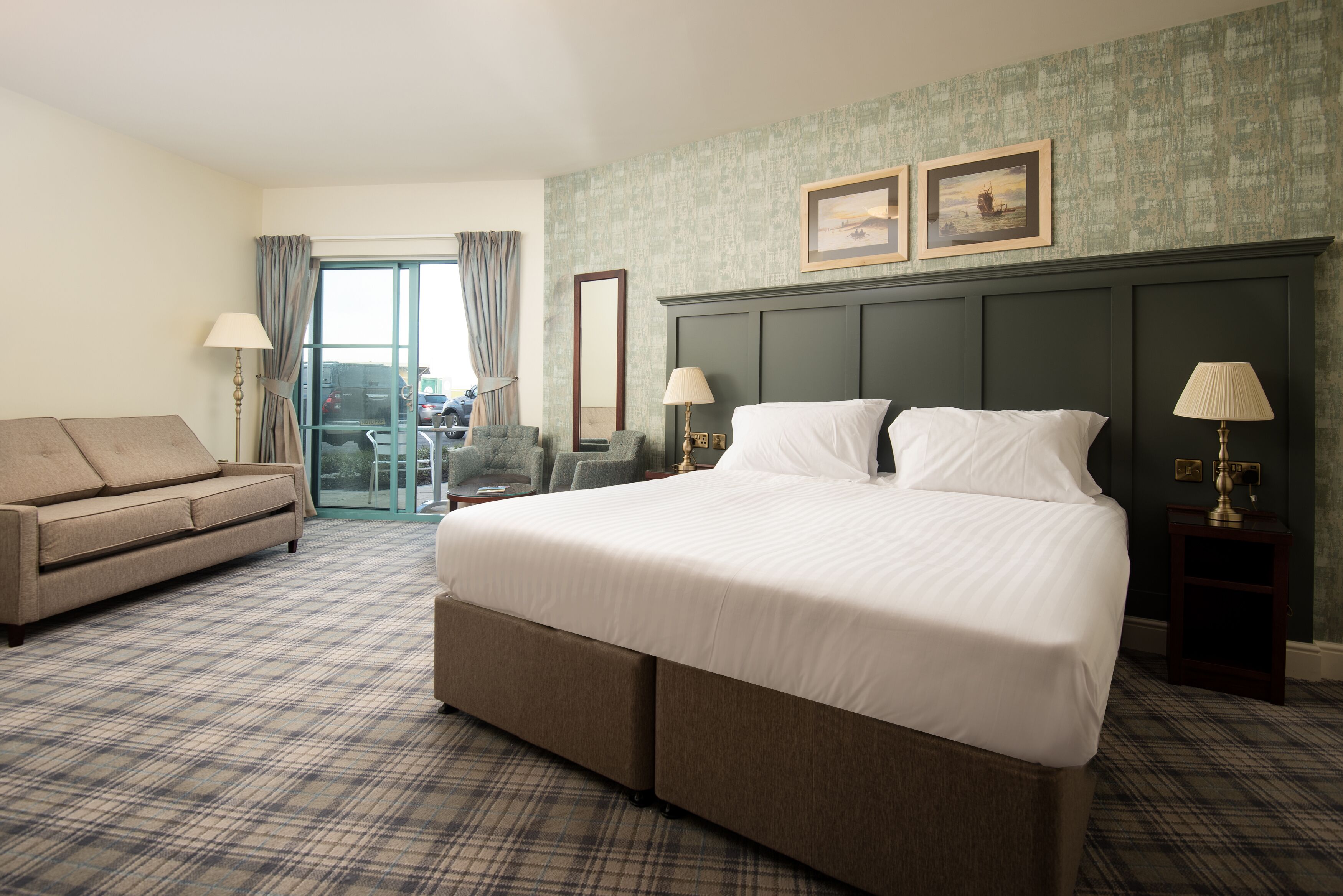 Habitación de hotel con una cama grande, un sofá, dos sillones, vistas al exterior y una pared verde con textura.