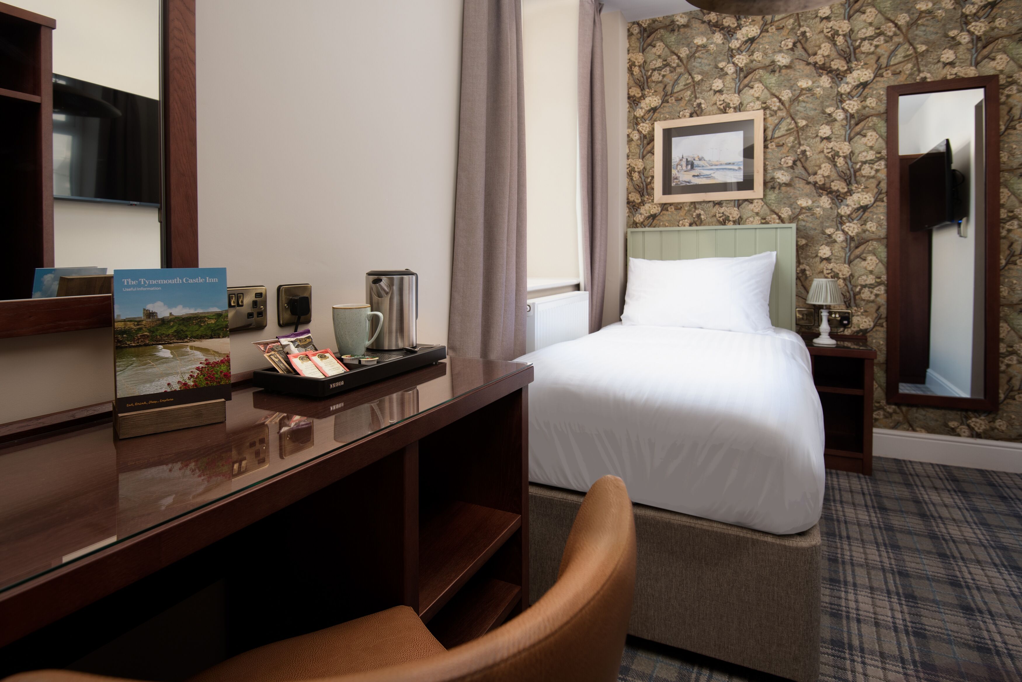 A hotel room with a bed, a desk with a computer, a chair, a nightstand with a lamp, and a framed picture on the wall.