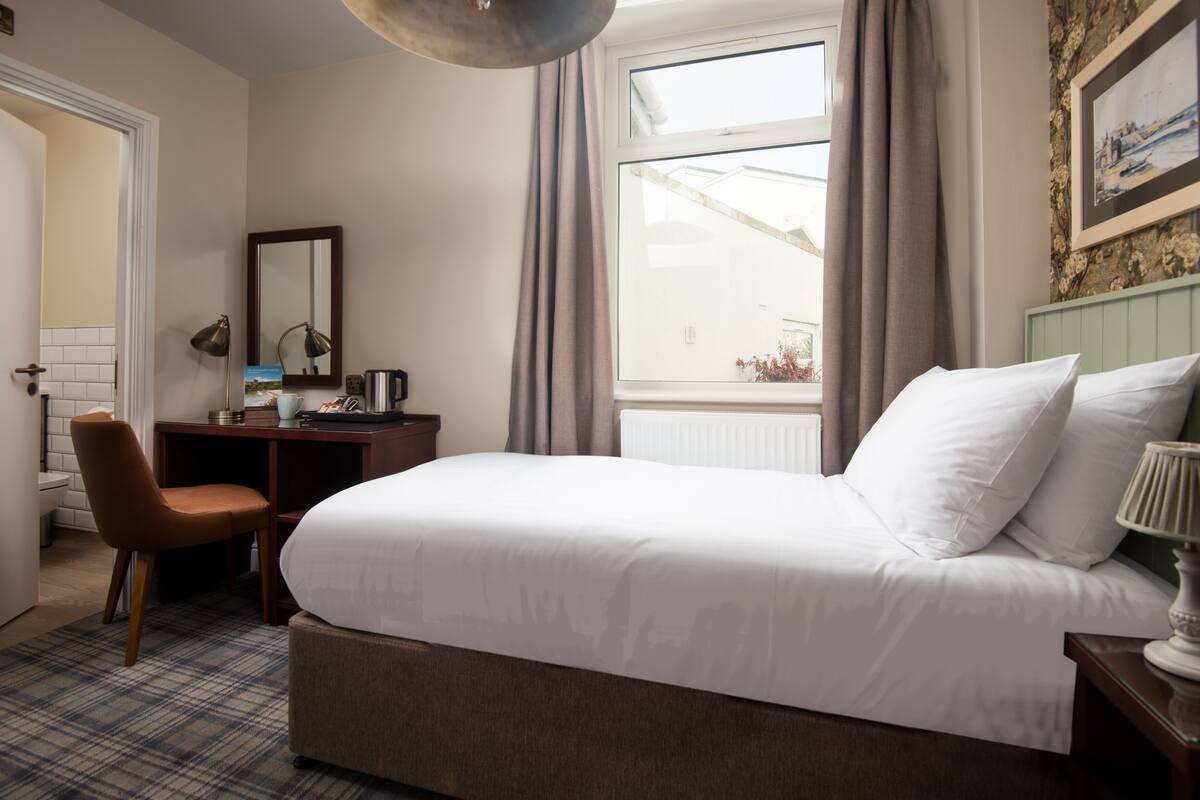 A hotel room with a large bed, a desk with a chair, a window with curtains, and a view of the outside.