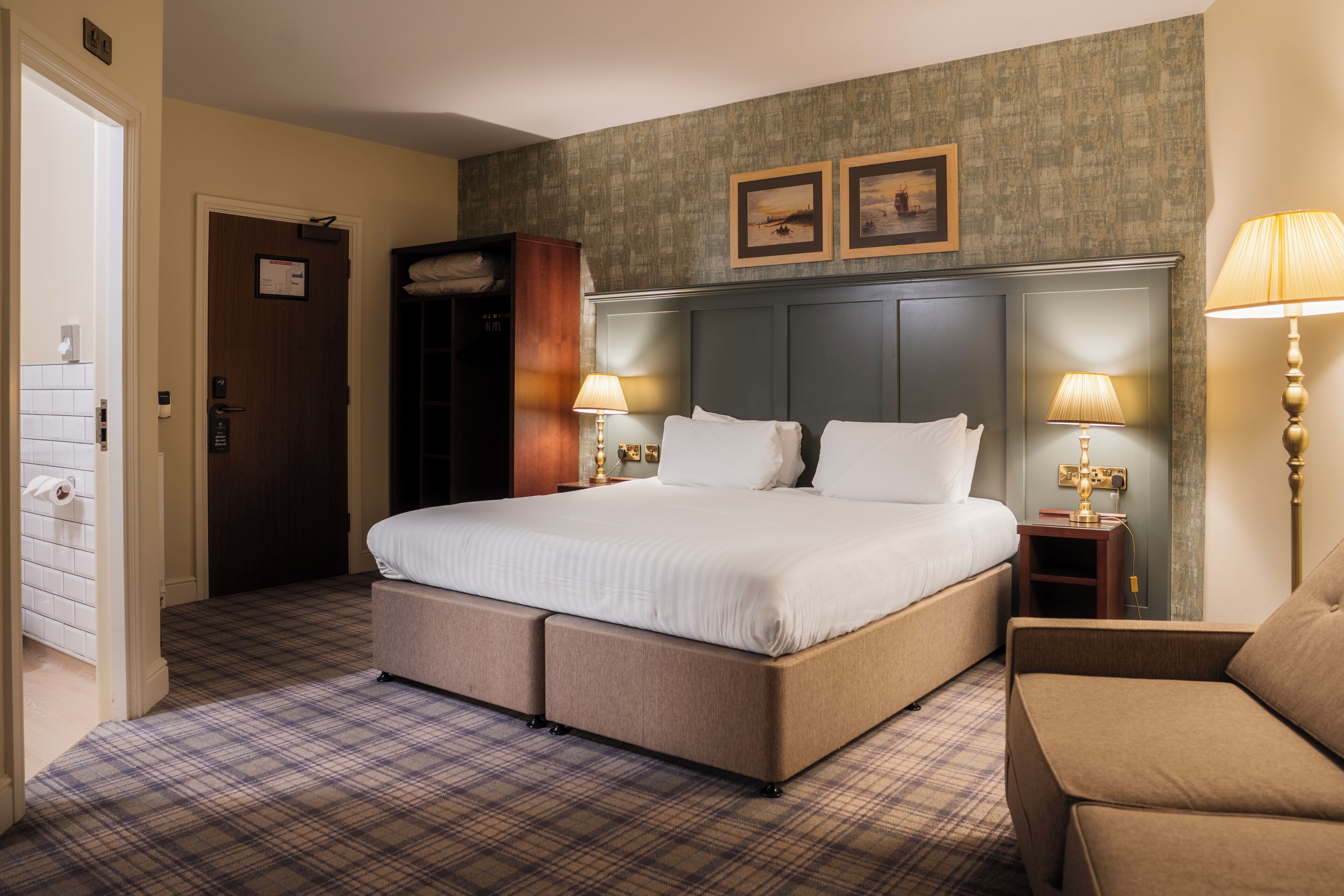 A hotel room with a large bed, two bedside tables with lamps, a wardrobe, and a plaid-patterned carpet.