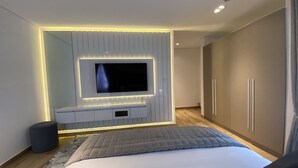 A modern bedroom with a large bed, a TV mounted on a wall with ambient lighting, a wardrobe, and a bedside table. - Mastermindsluxuryliving (Lagos)