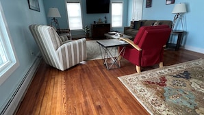 Una sala de estar con piso de madera, un sillón a rayas, una silla roja, un sofá, un televisor montado en la pared y un tapete estampado.