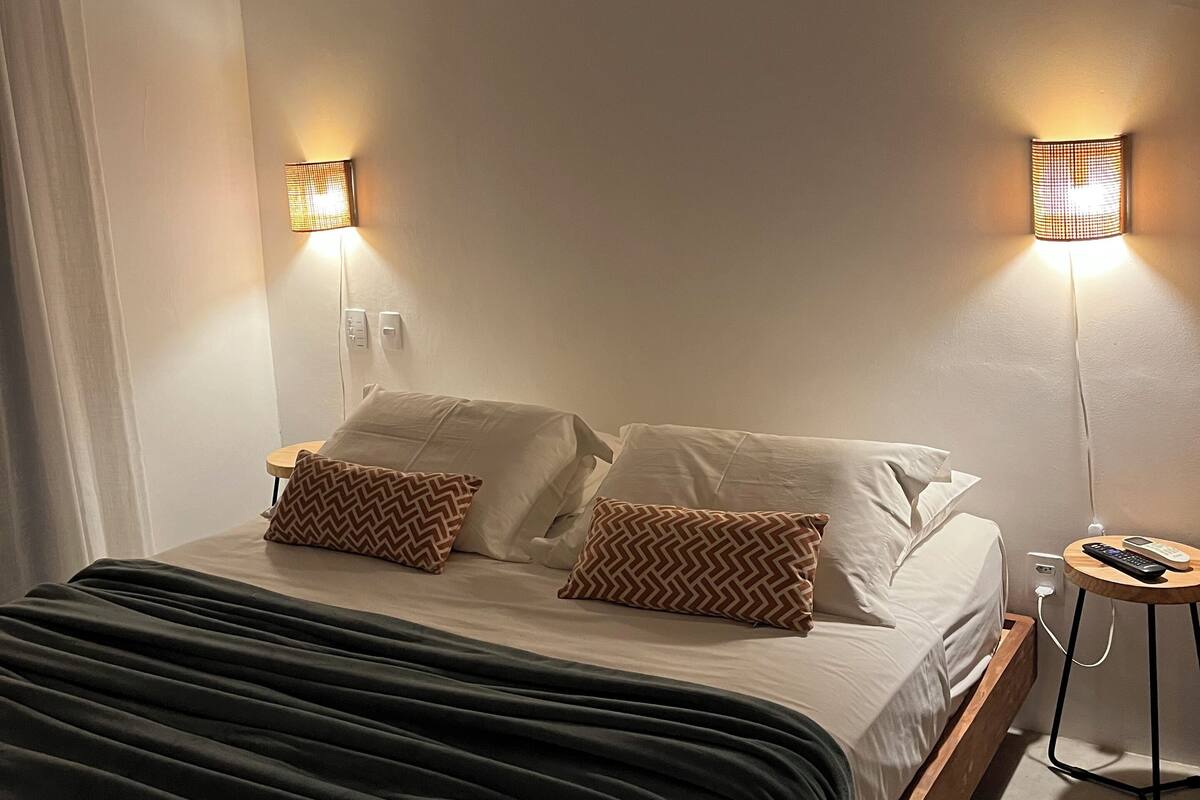 A neatly made bed with white linens and two patterned pillows, a bedside table with a remote control, and two wall-mounted lamps providing soft lighting.