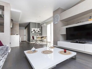 A modern living room with a white sofa, a dining area with a table and chairs, a large flat-screen TV, and a kitchen in the background. - Apartment Edison by Interhome (Peñiscola)