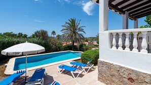 Zona de piscina con sillones reclinables azules y un paraguas blanco, rodeada de follaje y un cielo azul despejado.