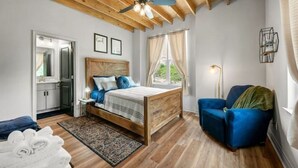 A bedroom with a wooden bed, a blue armchair, a nightstand, a mirror, and a window with curtains. - 5-Star 2BR: Clean, Comfy, SuperHost Hospitality (Bryan)