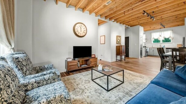 A spacious living room with a television, sofa, and coffee table. A kitchen area is visible in the background. - 5-Star 2BR: Clean, Comfy, SuperHost Hospitality (Bryan)