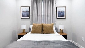 Una cama bien hecha con dos almohadas, una mesita de noche con una lámpara, y dos cuadros en la pared.