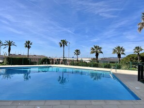 A rectangular swimming pool with a clear blue water surface, surrounded by a low green fence, palm trees, and a clear sky. - Bright studio just a stone's throw from the sea (Saint-Raphaël)