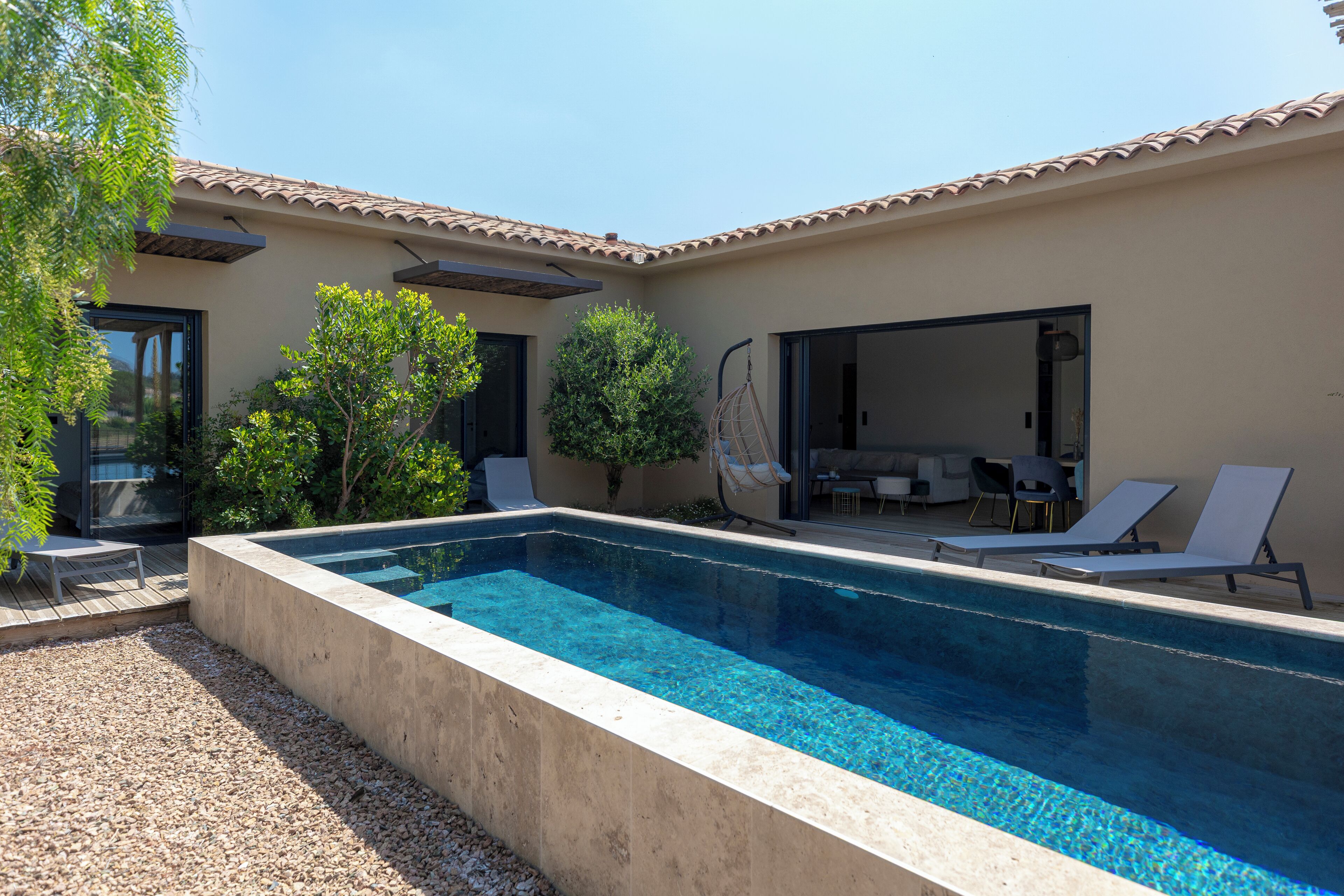 Una piscina rectangular con agua azul cristalina, rodeada por un muro color beige y un techo con azulejos. Hay sillas de descanso y una pequeña mesa cerca de la piscina, y una puerta que da a un espacio al aire libre.