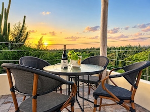 Une table ronde entourée de quatre chaises, une bouteille de vin et une plante en pot sur un balcon donnant sur un paysage de cactus et un coucher de soleil.