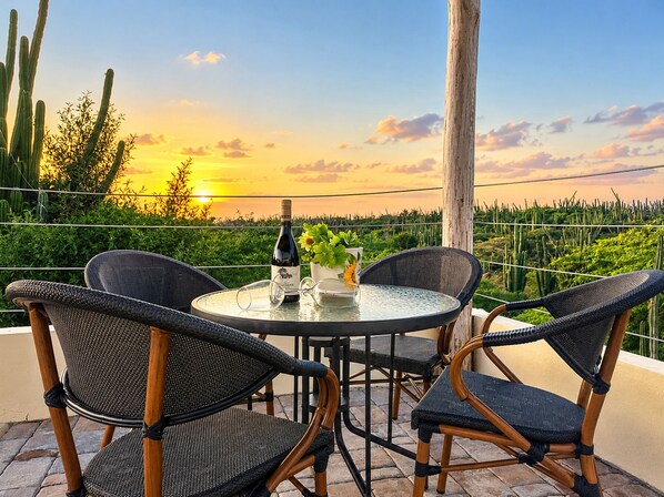 A round table with four chairs, a bottle of wine, and a potted plant on a balcony overlooking a landscape with cacti and a sunset.