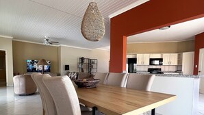 Un comedor amplio con una mesa de madera y sillas acolchadas, una lámpara colgante de fibra natural, y una cocina visible a través de un vano abierto.
