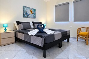 Un letto rifatto con cura, un asciugamano disposto a forma di cuore, un comodino con una lampada e una sedia con un cuscino.