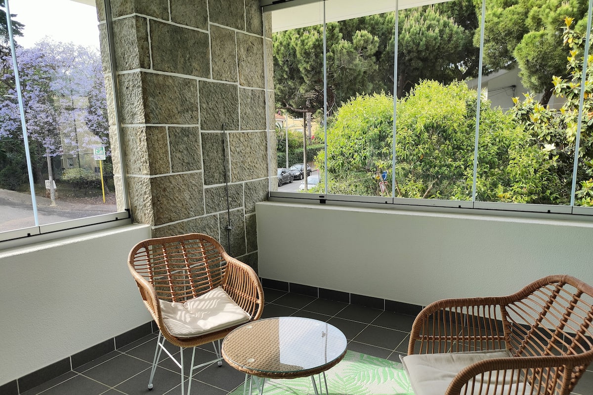 Una habitación con pared de piedra, ventana con vistas a árboles y coches aparcados, dos sillones de mimbre, una mesita redonda y una alfombra verde estampada.