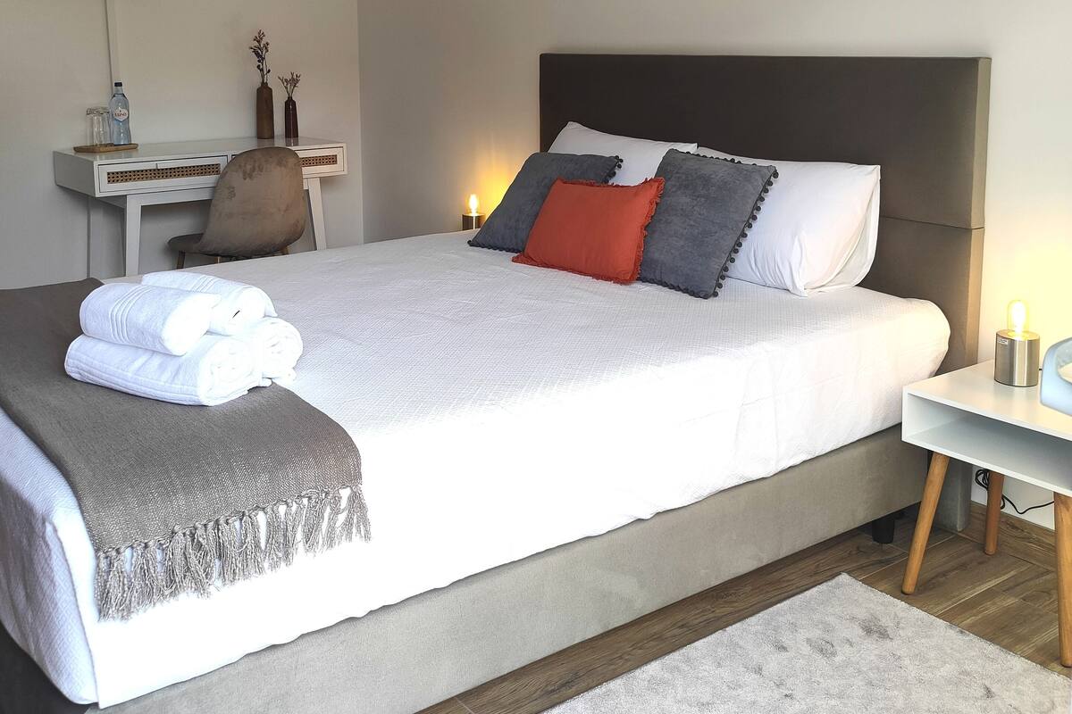 Una cama bien tendida con ropa blanca, un plaid gris y una mesita de noche con lámpara.
