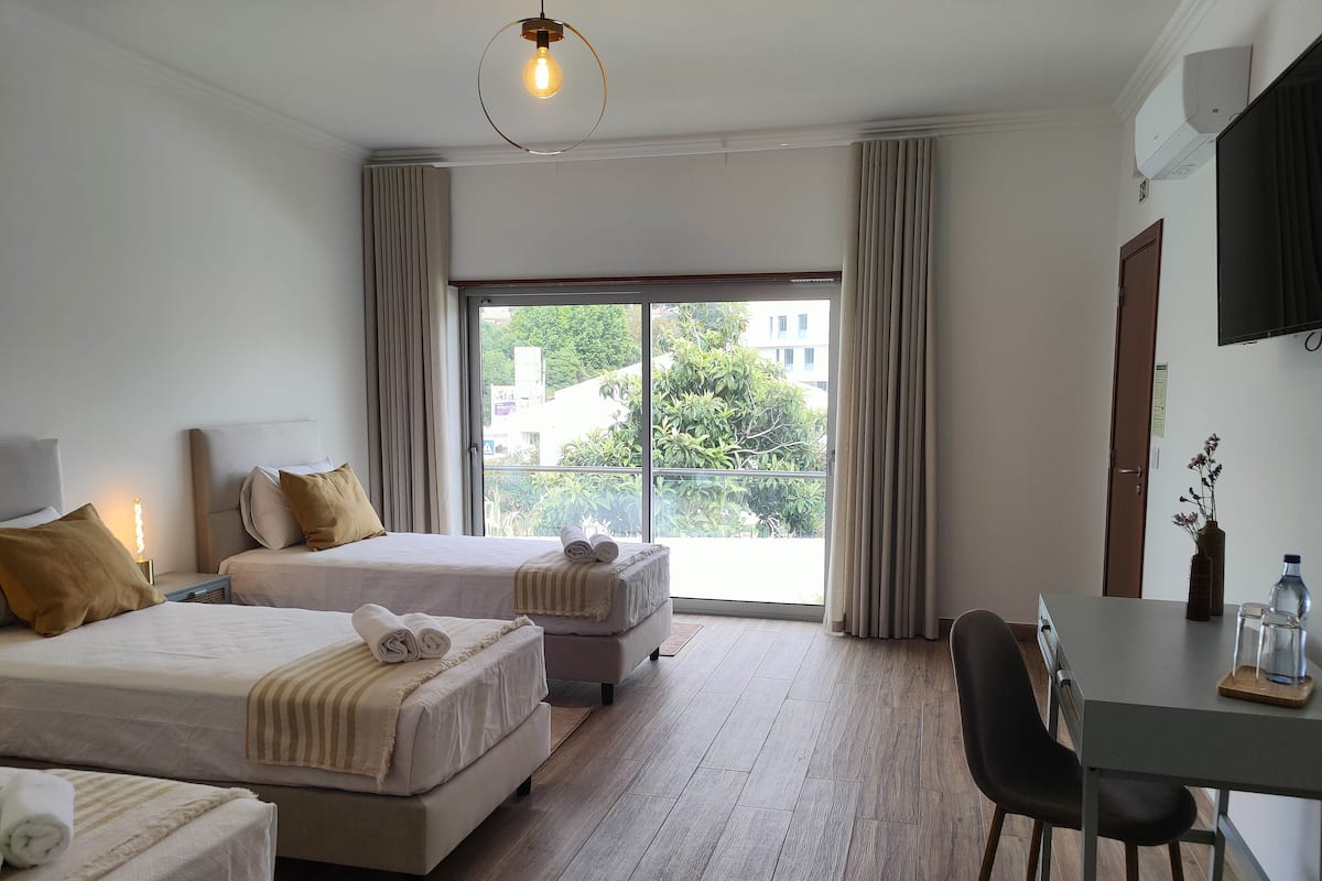 Habitación de hotel con dos camas, un escritorio, una silla, un televisor y un ventanal con vistas a edificios y zonas verdes.