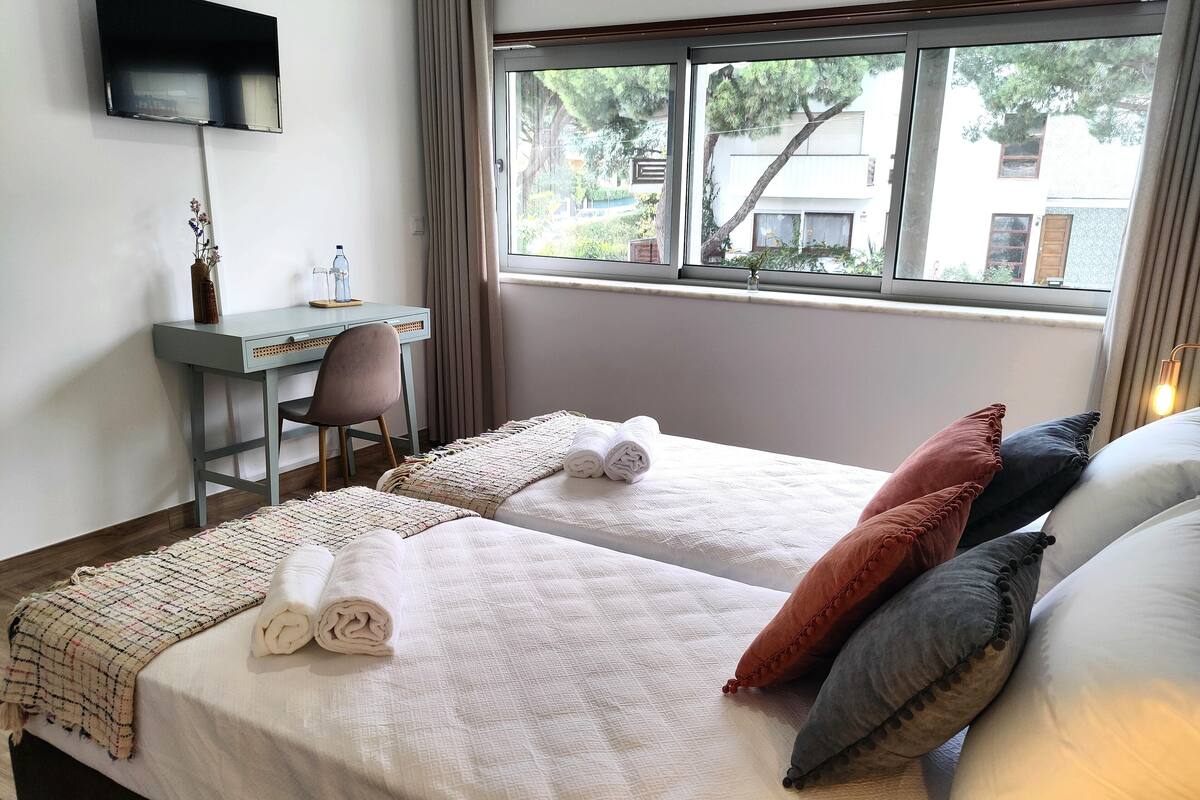 Habitación de hotel con dos camas, un escritorio, una silla, un televisor y una ventana con vistas a edificios y árboles.