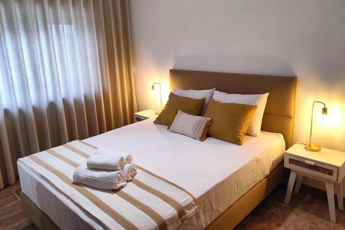 Una cama bien hecha con ropa de cama blanca y a rayas, dos lámparas de noche y una ventana con cortinas.