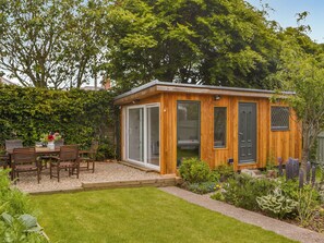 A wooden cabin with large windows, a patio with a table and chairs, and a garden with various plants.