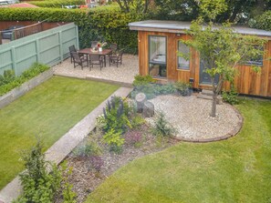 A backyard with a wooden deck, a dining table with chairs, a small shed, and a garden with various plants and a tree.