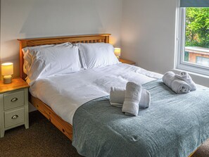 A neatly made bed with pillows and a blanket, a bedside lamp, a nightstand with a drawer, and a window with a view of greenery.