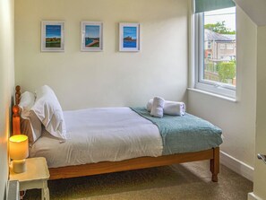 Un lit simple avec une literie blanche, un plaid turquoise et deux serviettes enroulées. Trois tableaux encadrés ornent le mur, ainsi qu’une lampe de chevet et une fenêtre donnant sur les bâtiments environnants.