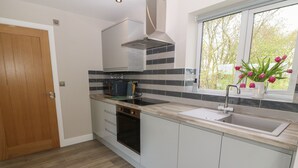 A modern kitchen with a built-in oven, a stainless steel range hood, a double sink, and a window with a view of trees. - Bwthyn Cae'r Efail (Rhosgoch)