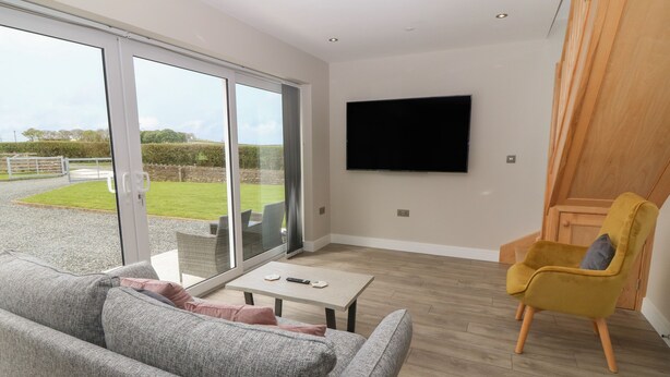 A modern living room with a grey sofa, a yellow armchair, a flat-screen TV, and a sliding glass door leading to a garden. - Bwthyn Cae'r Efail (Rhosgoch)
