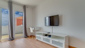 A modern living room with a flat-screen TV mounted on the wall, a white TV stand with drawers, a white chair, and a large sliding glass door leading to an outdoor area. - Esfera I (Santa Cruz)