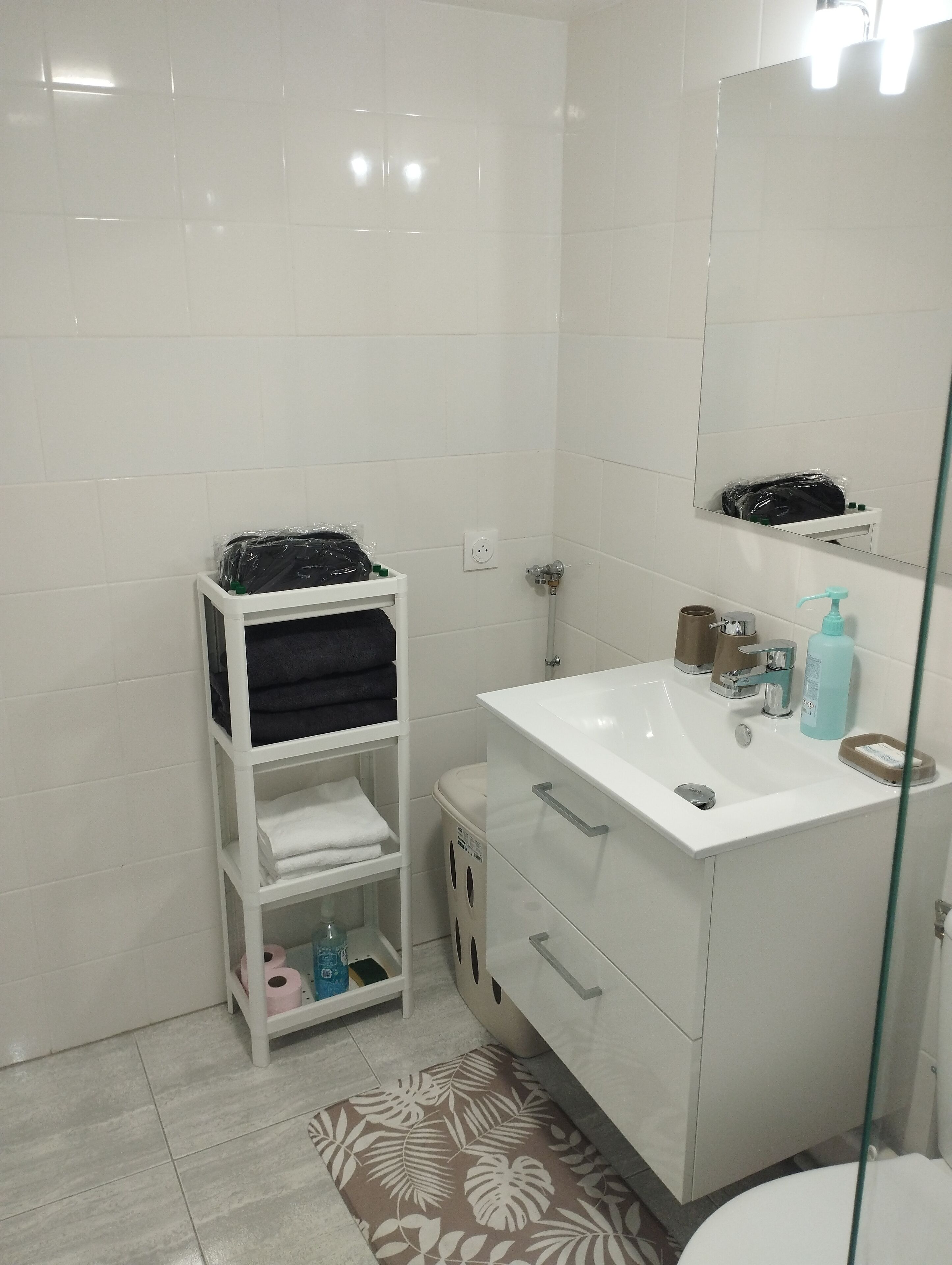 Une salle de bain avec des carreaux blancs, un lavabo blanc avec un miroir, une douche en verre et une étagère blanche où sont posés des serviettes et des articles de toilette.