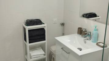 Une salle de bain avec des carreaux blancs, un lavabo blanc avec un miroir, une douche en verre et une étagère blanche où sont posés des serviettes et des articles de toilette.