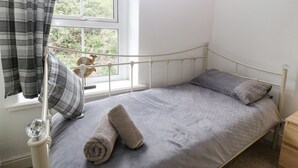 A single bed with a metal frame, grey bedding, and a window with curtains. - Pebblestone Cottage (Llandudno)