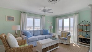 Un salon avec un canapé bleu clair, un pouf blanc, des fauteuils en osier et une bibliothèque.