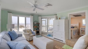 Une chambre donnant sur la plage, avec un canapé blanc, un fauteuil en osier, une armoire blanche et une vue sur la plage à travers de grandes fenêtres.