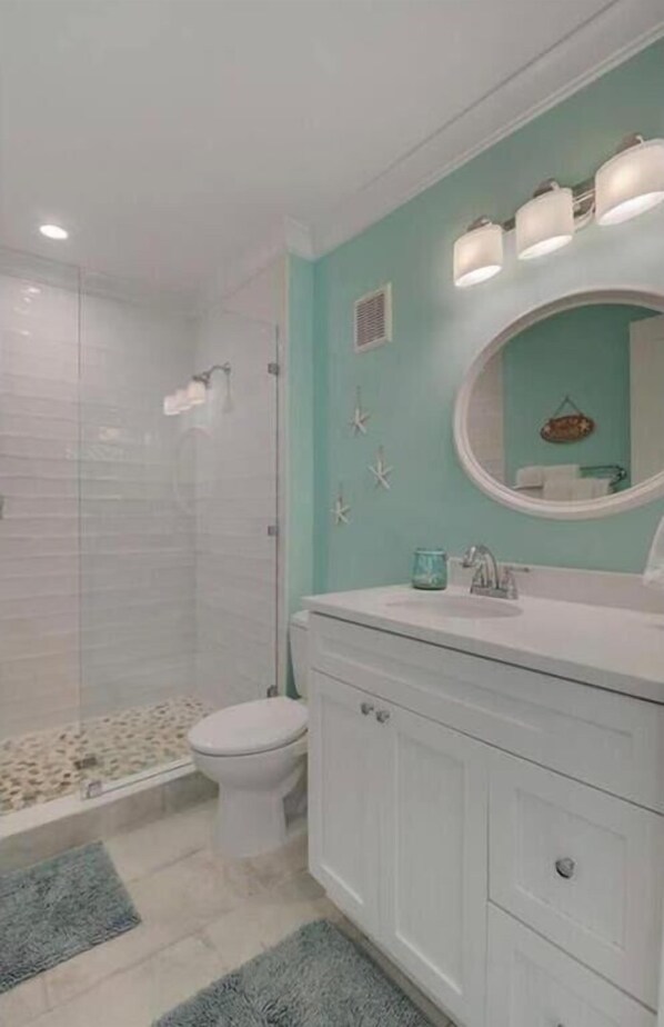 A bathroom with a glass-enclosed shower, a white toilet, a vanity with a round mirror, and a teal accent wall.