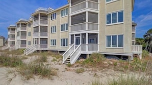 Un immeuble à plusieurs étages situé en bord de mer, doté de balcons et d’un escalier menant à l’entrée, avec une plage de sable au premier plan.