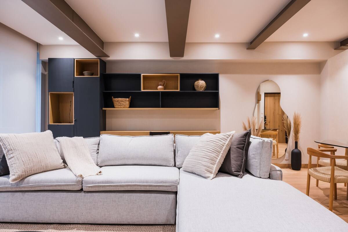 A modern living room with a grey sectional sofa, wooden shelves, and a dining area with a glass table and chairs.