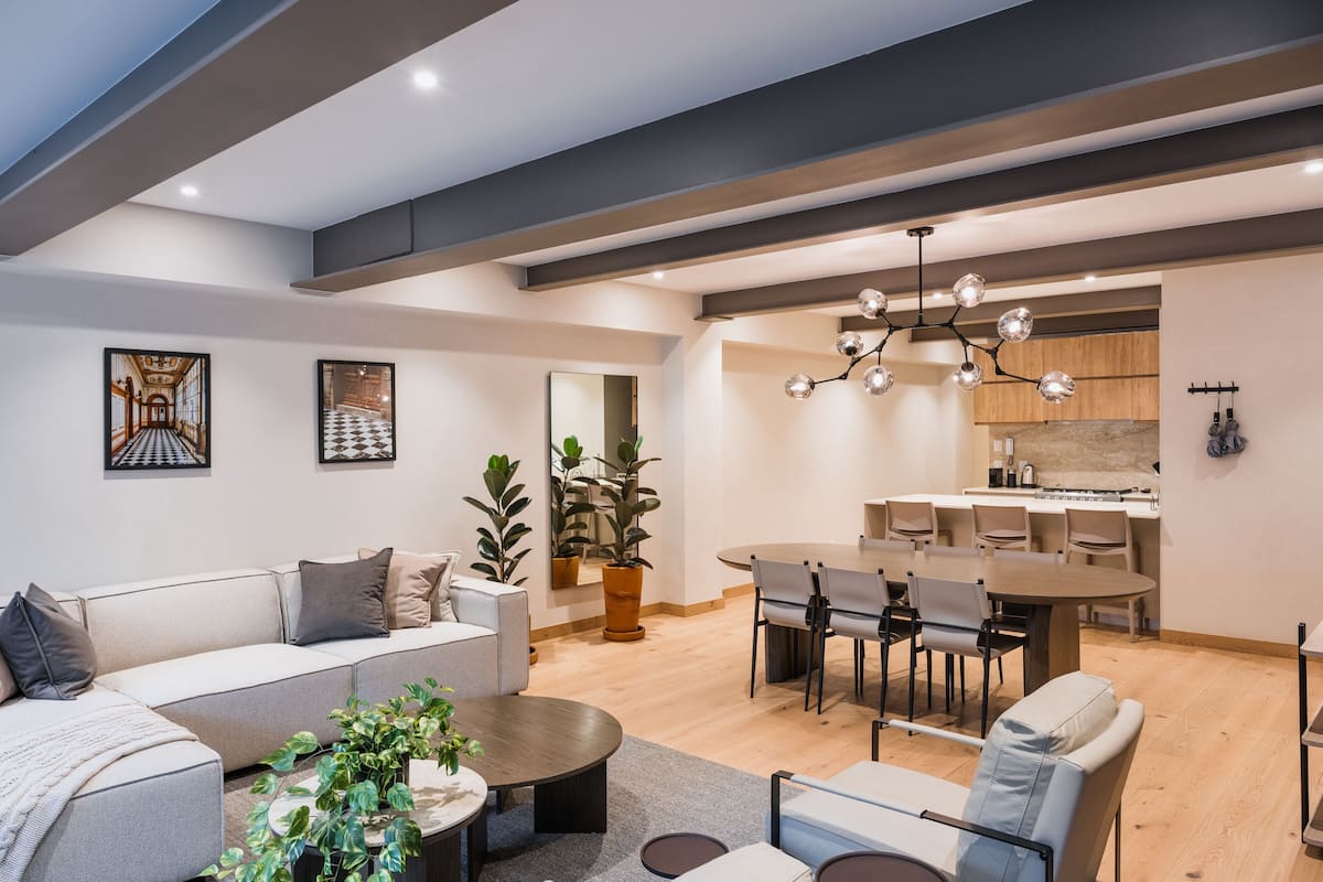 A modern living room with a sofa, coffee table, and dining area. There are potted plants and artwork on the walls.