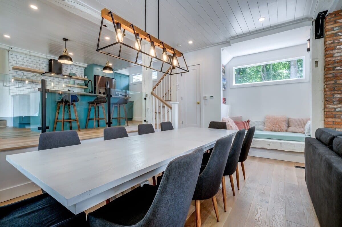 A modern kitchen with a central island, a dining area with a long table and chairs, and a cozy seating area with a sofa and cushions.