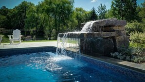 A swimming pool with a waterfall feature, surrounded by greenery and a white chair. - 3-bedroom house 915A Shefford mountain view (Bromont)